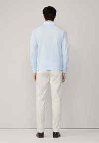 Svijetloplava košulja dugih rukava s dugmadima na manžetama, u kombinaciji s kremastim hlačama i tamnim cipelama, prikazana s leđa.