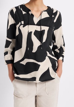 Kvinde iført en sort og beige bluse med abstrakt print og trekvartærmer samt cremefarvede bukser, hænderne i lommerne mod en hvid baggrund.