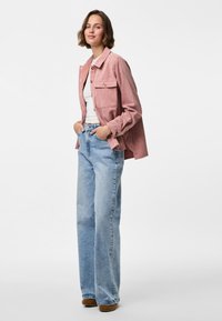 Roze corduroyjas met twee borstzakken, gecombineerd met lichtblauwe wijde jeans. Textuurstof en relaxte pasvorm, aangevuld met bruine schoenen.