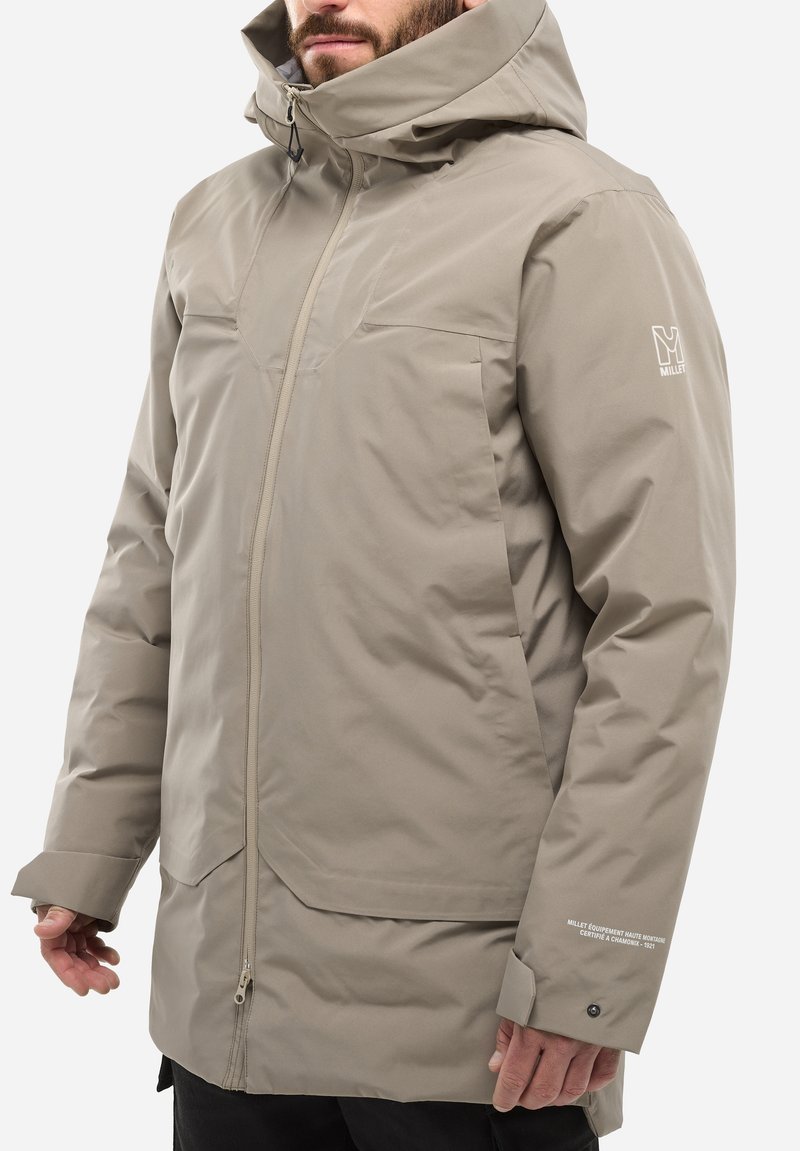 Veste isolée beige clair avec capuche, fermeture éclair à l'avant et poignets ajustables. S'agence avec un logo sur la manche et des éléments de design épurés.