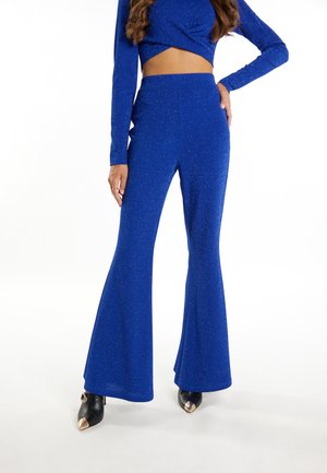 Blauwe glinsterende uitlopende broek met een soepele textuur, hoge taille en een aansluitend ontwerp, gecombineerd met zwarte puntige hakken met gouden accenten.