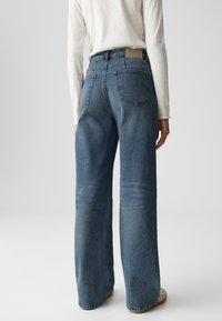 High-waisted, wide-leg jeans in medium blue denim, featuring a faded texture, double back pockets, and a beige waistband label.