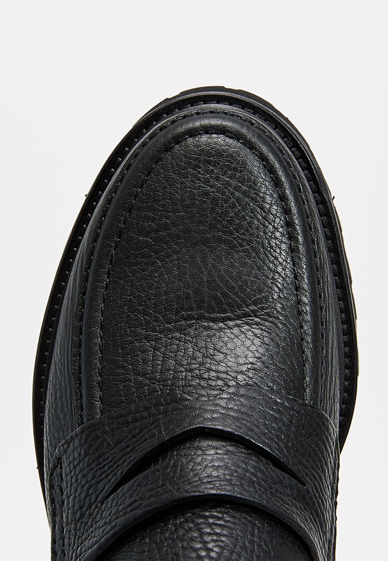 Schwarze Lederloafers mit strukturierter Oberfläche, abgerundeter Zehe und dekorativem Riemen über dem Obermaterial. Gesteppte Details entlang der Sohle.