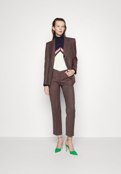 Καφέ παγιέτα κοστούμι με κομψή εφαρμογή, τοπ σε λευκό και ναυτικό μπλε χρώμα με μπλοκαρισμένες αποχρώσεις και πράσινα στιλέτο πέδιλα. Μοντέλο στέκεται μπροστά σε ουδέτερο φόντο.