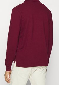 Bordeaux långärmad polo med krage, sidslitsar och en mjuk textur, designad för komfort och rörelsefrihet.