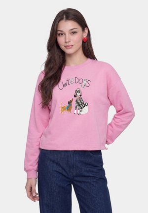 Jonge vrouw met lang donker haar die een roze sweatshirt draagt met twee honden en de tekst "Cute Dogs" en oorbellen in de vorm van een rood hart.