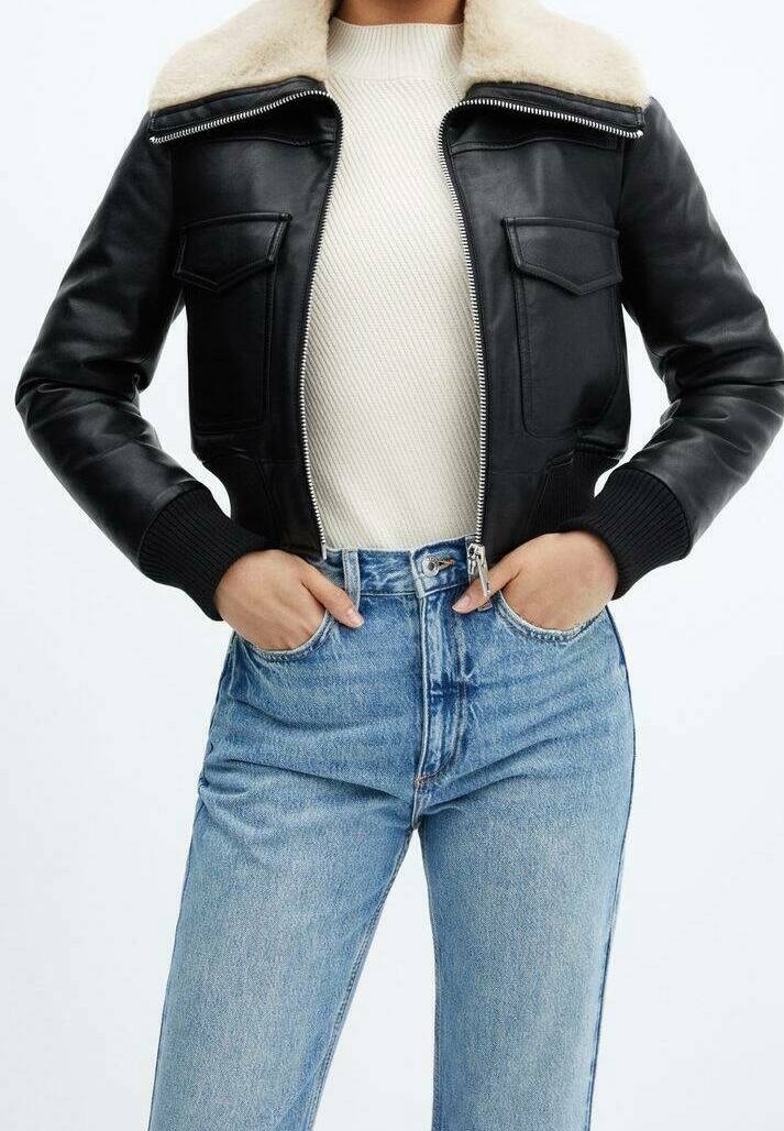 Veste en cuir noir avec col en shearling portée sur un pull côtelé blanc et un jean taille haute bleu clair, les mains dans les poches.