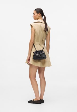 Woman in beige short-sleeve dress with belted waist, black woven flats, and black quilted handbag with chain strap over shoulder, facing sideways.