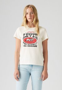 Kremveida kokvilnas T-krekls ar sarkanu un melnu grafiku, kurā redzami zirgi un teksts "LEVI'S THE ORIGINAL". Īsās piedurknes, taisns piegriezums.