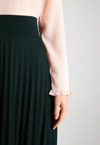 Pitkähiha, vaaleanpunainen pusero, jossa on röyhelöhihoja, yhdistetty tummanvihreään laskostettuun hameeseen, joka korostaa pinnan sileitä tekstuureja ja kontrastoivia värejä.