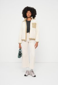 Giacca in pile color avorio con rifiniture beige e taschino sul petto, abbinata a pantaloni a zampa color crema chiaro e sneakers grigie. Tiene in mano una borsa verde.