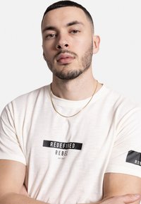 T-shirt beige clair avec un design graphique noir disant "REDEFINED REBEL EST. 2017," présentant un col rond et des manches courtes.