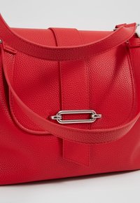 Sac à main en cuir rouge texturé avec rabat, fermoir rectangulaire argenté et large bandoulière sur fond clair.