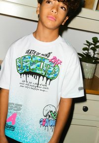 Weißes Baumwoll-T-Shirt mit buntem Grafikdruck, schwarzen Tropfen, einem Totenkopfmotiv und blauen Farbverlauf-Details. Kurze Ärmel und Rundhalsausschnitt.