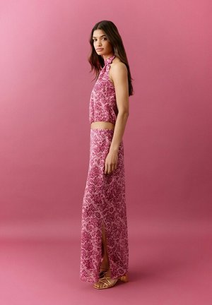 Une femme se tient devant un fond rose, portant un haut à col licou sans manches à motifs floraux et une jupe longue assortie avec une fente latérale, combinés à des sandales dorées.