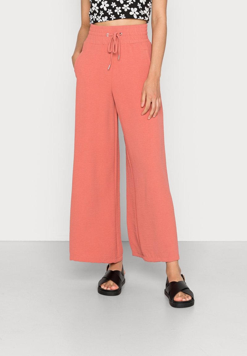 Korallfärgade vida byxor med elastisk midja och snörningsdetalj. Bärs med svarta platta sandaler; bakgrunden är minimalistisk och vit.