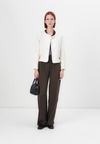 Veste blanche texturée courte avec fermeture à bouton, portée sur un haut à motifs. Pantalons marron à rayures verticales amples et petit sac à main noir.