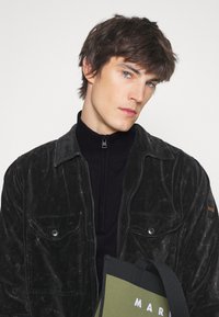 Schwarze gewachste Jacke mit Fronttaschen, über einem schwarzen hochkragen Pullover getragen. Enthält eine grüne Tragetasche mit weißer Schrift.