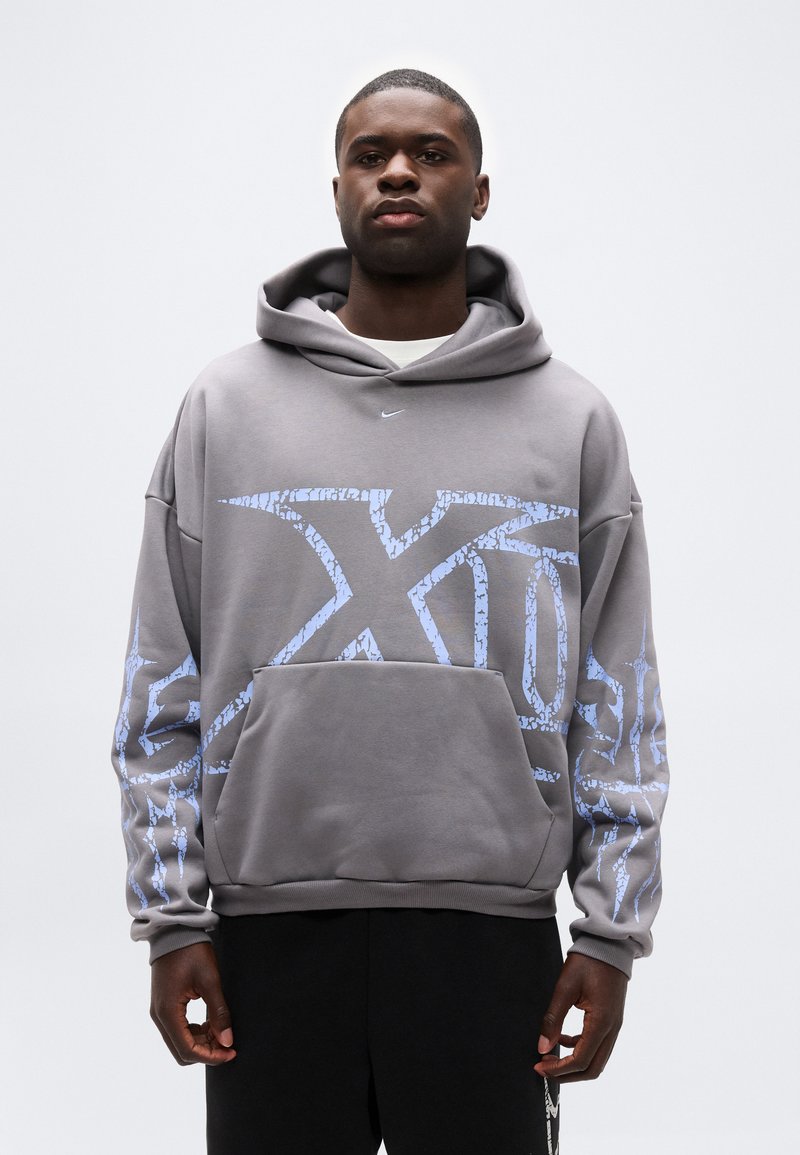 Mann trägt grauen Hoodie mit großem blauem abstraktem Muster und schwarze Hose, steht vor weißem Hintergrund.