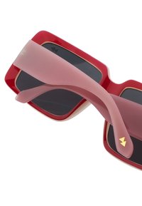 Červené sluneční brýle se černými skly. Mají růžovou průhlednou nožičku, zlaté akcenty a lesklý povrch. Vysoký kontrast designu.