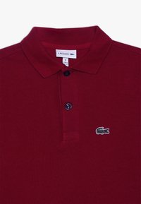 Rotes Baumwoll-Polohemd mit klassischem Kragen, zwei Knöpfen und gesticktem grünen Krokodil-Logo auf der linken Brust.