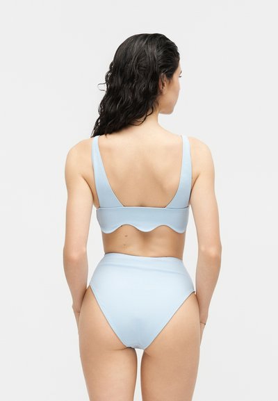 Femme aux longs cheveux foncés portant un bikini taille haute bleu clair avec un haut à bord festonné, vue de dos sur un fond uni.