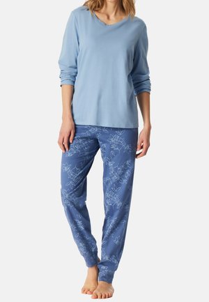 Woman wearing light blue long-sleeve top and blue pants with white floral patterns, standing barefoot on a plain background.