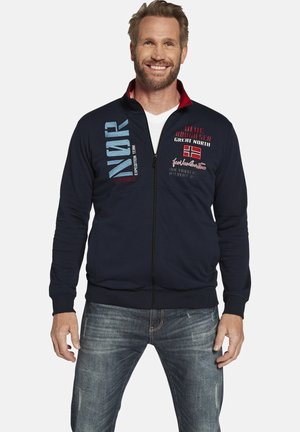 Navy zip-up jacket made of cotton blend, featuring red accents, printed text, and a relaxed fit. Worn with light denim jeans.