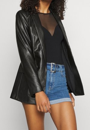 Nainen pukeutuneena mustaan nahkatakkiin, mustaan läpikuultavaan toppiin ja korkeavyötäröisiin sinisiin farkkushortseihin, joissa on käännetyt lahkeet ja hopeanvärinen solkivyö.