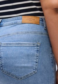 Φωτεινά γαλάζια τζιν με λεπτομέρεια πίσω τσέπης και καφέ δερμάτινη ετικέτα бренда στη ζώνη με ραφές διακόσμησης.