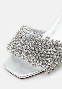 Sandale à talons hauts argentée avec un bout carré, ornée d'un amas de strass multiformes sur le dessus. Texture lisse et métallique.