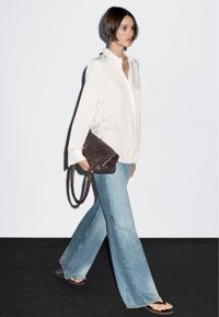 White button-up shirt, light blue flared jeans, and brown woven handbag. Model wears black flip-flops. Simple, clean design and textures.