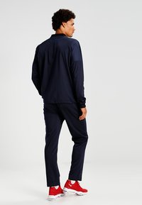Tuta sportiva navy con maniche lunghe, colletto alto e pantaloni abbinati. Presenta scarpe da ginnastica rosse e una texture liscia e opaca.