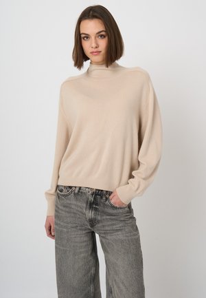Junge Frau mit kurzen braunen Haaren, die einen beigen Rollkragenpullover und ausgewaschene graue Jeans trägt, eine Hand in der Tasche, neutraler Hintergrund.