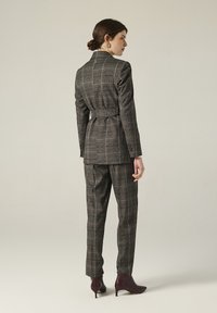 Donna che indossa un abito a trama gessata grigia con pantaloni su misura e un blazer abbinato con cintura, in piedi di profilo con i capelli raccolti in un basso chignon e tacchi bordeaux.