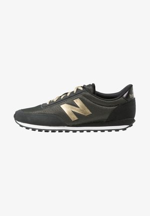 Black sneaker with a textured upper, gold accents, and a prominent silver "N" logo. Features a white midsole and black rubber outsole.