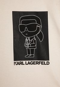 Gaišas t-krekls ar melnu zīmējumu, kurā attēlots figūra saulesbrillēs un uzvalkā, ar režģa fona un tekstu "KARL LAGERFELD" zem tā.