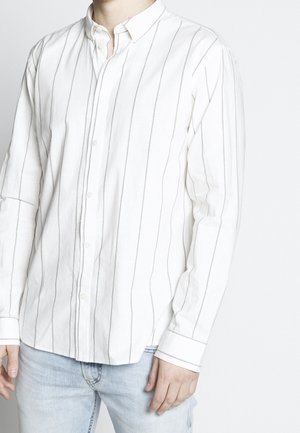 Mężczyzna ubrany w białą koszulę na guziki z długim rękawem, z cienkimi pionowymi szarymi paskami oraz jasnoniebieskie dżinsy, stojący na jednolitym tle.