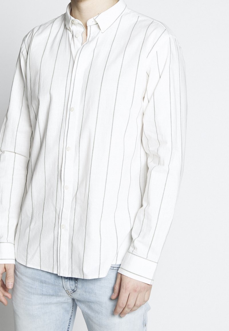 Homme portant une chemise blanche à manches longues avec de fines rayures verticales grises et un jean bleu clair, debout devant un fond uni.