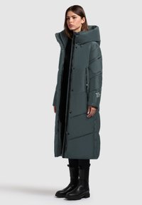 Padded dugi kaput u tamno tirkiznoj boji s visokim ovratnikom, višestrukim džepovima i kopčama na dugme. Uparen s crnim gležnjačama. Jednostavan dizajn.