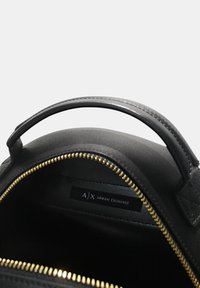 Svart håndveske med teksturert overflate, gullglidelås og kort hank. Innvendig har den en etikett med "A/X ARMANI EXCHANGE."