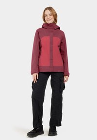 Veste bordeaux avec un panneau rouge contrastant, dotée d'une capuche, de poches zippées et d'un design ajusté. Associée à un pantalon cargo noir et des chaussures.