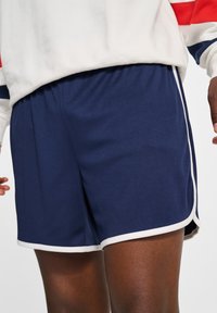 Osoba ubrana w granatowe sportowe szorty z białym obramowaniem oraz białą bluzę z czerwono-niebieskimi paskami na rękawach.