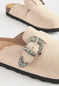 Des chaussures slip-on en suede rose clair avec une semelle en caoutchouc noir et un détail de boucle en argent ornée. Bout rond et texture lisse.