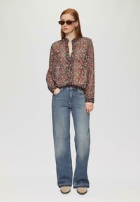 Blouse noire en tissu transparent à motif floral, avec un col froncé, fermeture à boutons et coupe ample, associée à un jean bleu clair évasé et des chaussures beiges.