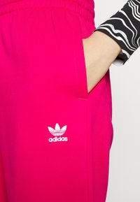 Růžové tepláky s elastickým pasem, na kterých je bílé logo Adidas. Zblízka je vidět boční kapsa a hladká textura látky.