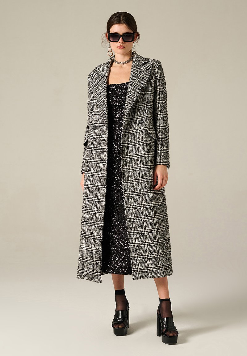 Cappotto lungo a quadri in bianco e nero, con un design sartoriale, doppio petto e rever a punta. Indossato sopra un abito nero di paillettes.