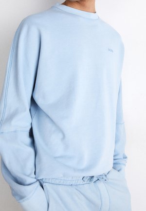 Man som bär en ljusblå långärmad sweatshirt och matchande dragsko byxor, står med händerna i fickorna, med logotypen "BOSS" på bröstet.