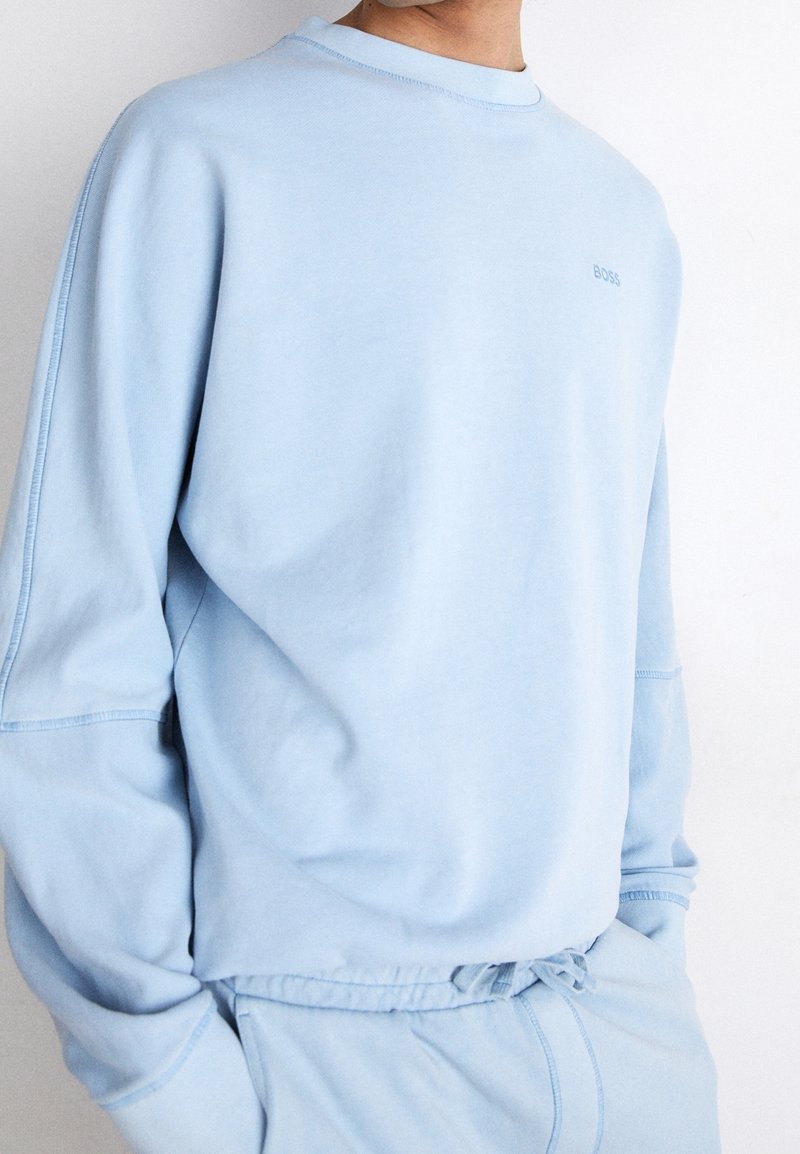Man som bär en ljusblå långärmad sweatshirt och matchande dragsko byxor, står med händerna i fickorna, med logotypen "BOSS" på bröstet.