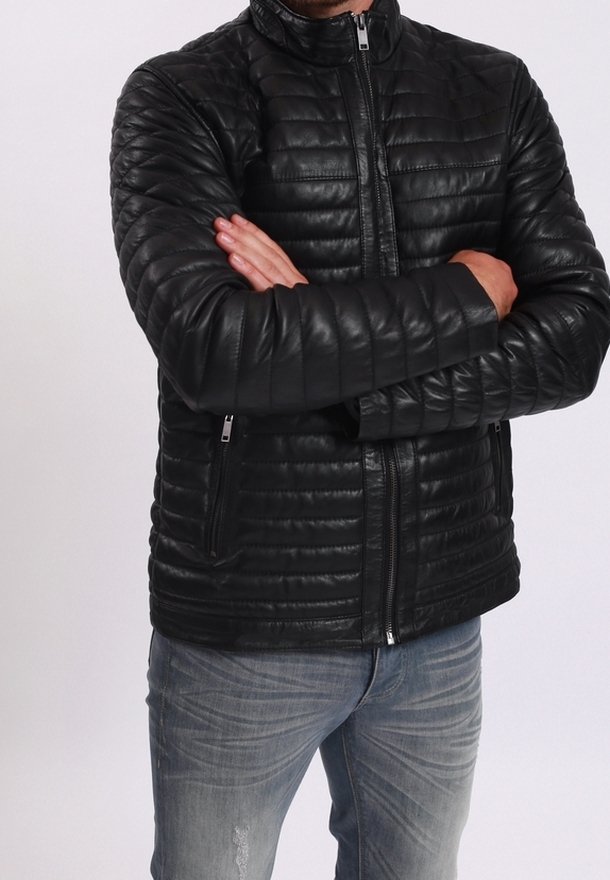 Homme bras croisés portant une veste matelassée noire zippée et un jean bleu délavé sur un fond blanc uni.